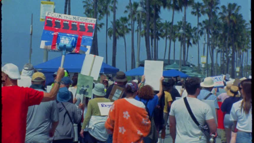 Vintage-style handheld footage follows a crowd of protestors marching down a palm-tree-lined street in California