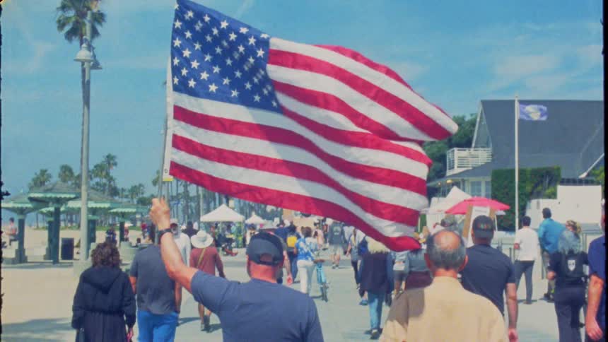 Vintage-style slow-motion footage shows a man proudly waving a large American flag while leading a crowd of people down a sunny beach boardwalk