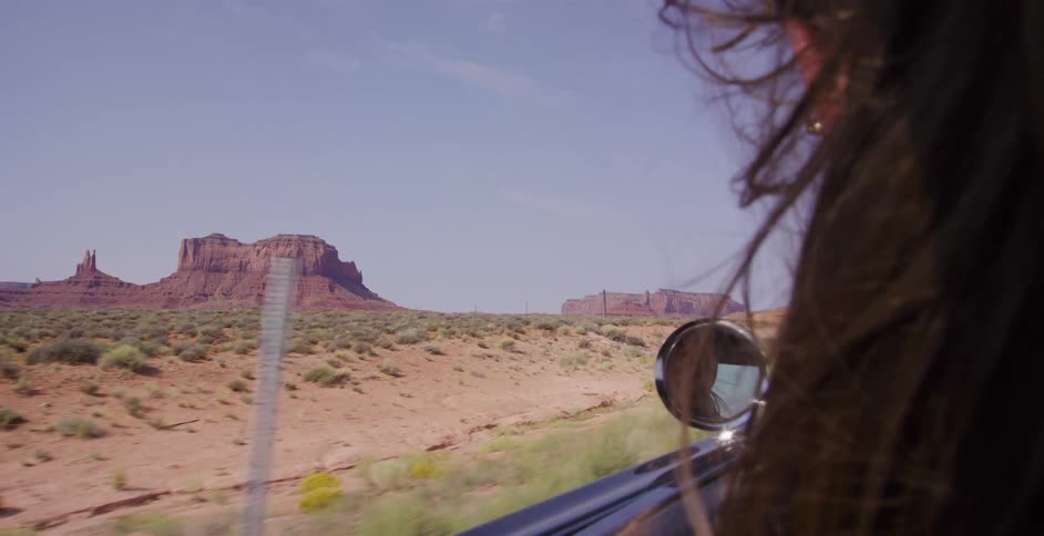 A woman with her hair blowing in the wind looks out a car window at the scenic buttes of Monument Valley