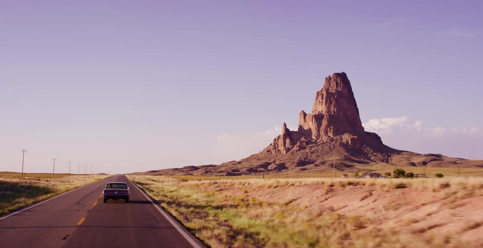 A classic car travels down an empty desert highway towards a large rock formation at sunset