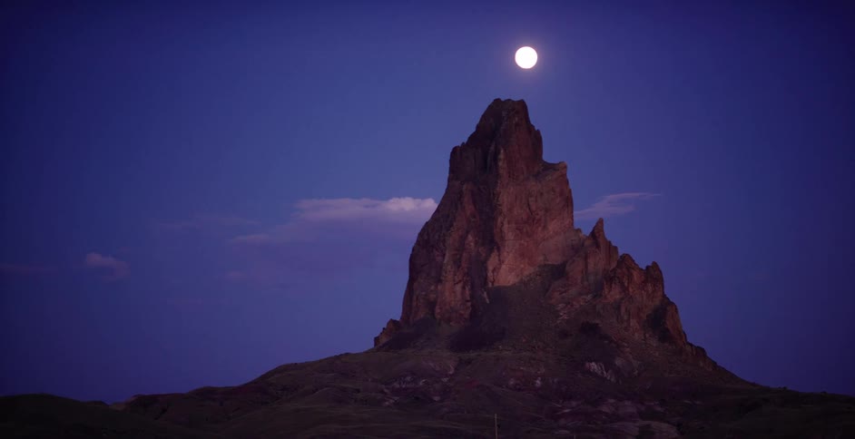 A full moon shines brightly in the deep blue night sky over a dramatic desert rock formation as clouds drift by
