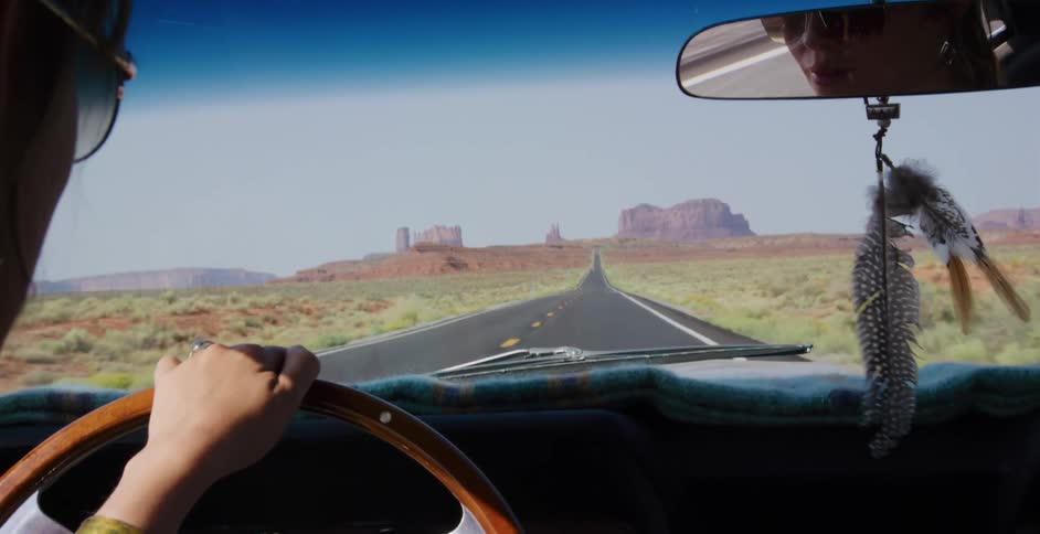 Over-the-shoulder view of a woman driving a vintage car down a long desert highway toward the buttes of Monument Valley