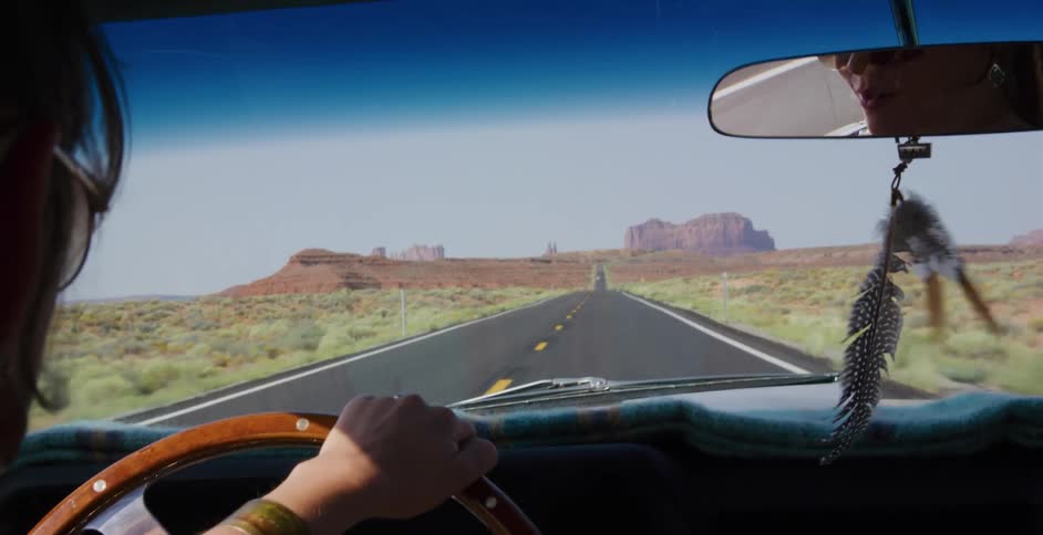 An over-the-shoulder shot captures the view from inside a vintage car driving towards the iconic Monument Valley