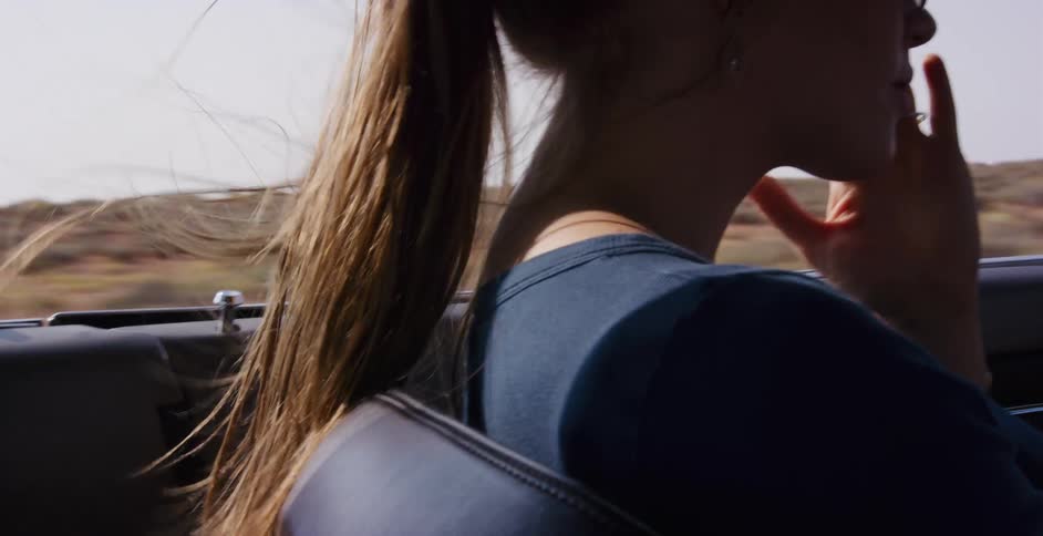 A woman's ponytail blows in the wind as she drives a car on a road trip through a desert landscape