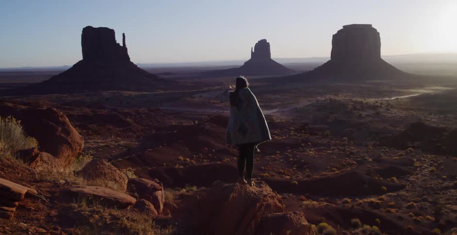A woman walks to the edge of a cliff overlooking Monument Valley at sunrise and unfurls a blanket that billows in the wind