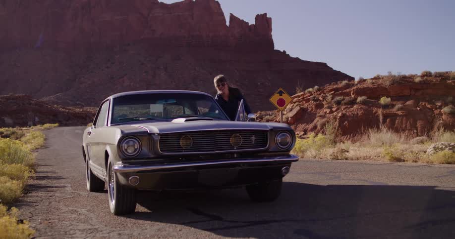A woman gets out of a vintage Ford Mustang parked on a remote desert road with red rock canyons in the background as the camera slowly pushes in
