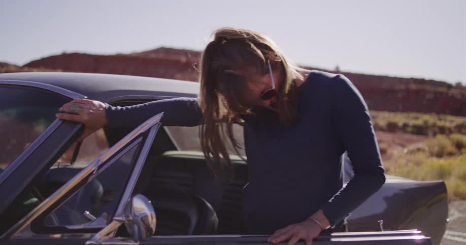 A carefree woman in sunglasses exits her classic car and playfully runs down a desert road in slow motion