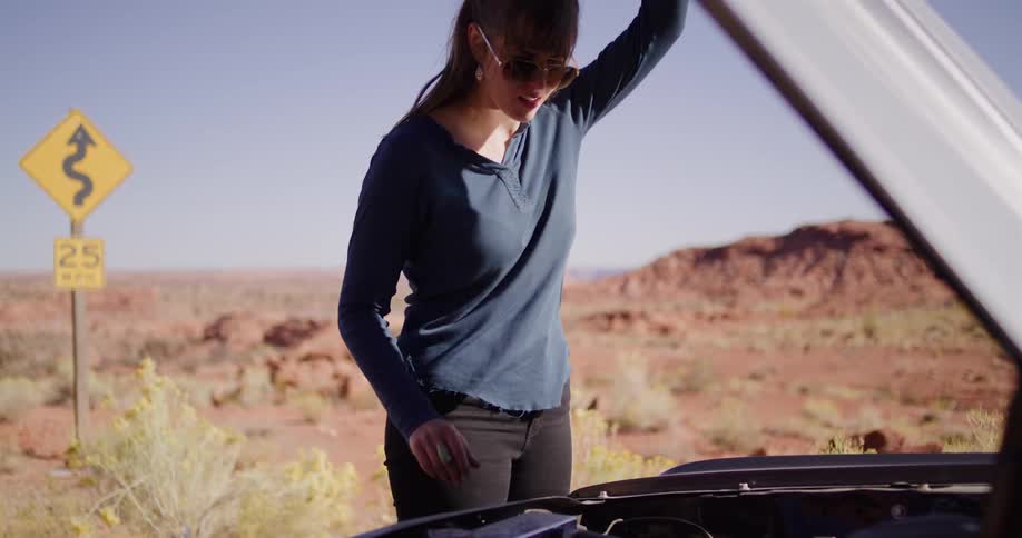 A frustrated woman gives up trying to fix her broken-down car and slams the hood shut on a deserted desert road