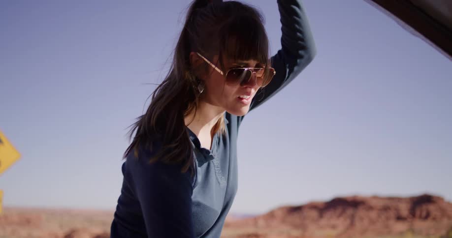 A frustrated woman in sunglasses inspects the engine of her broken down car on the side of a desert highway