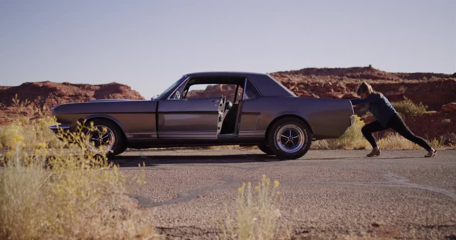 A determined woman push-starts her broken-down classic muscle car on a remote desert highway