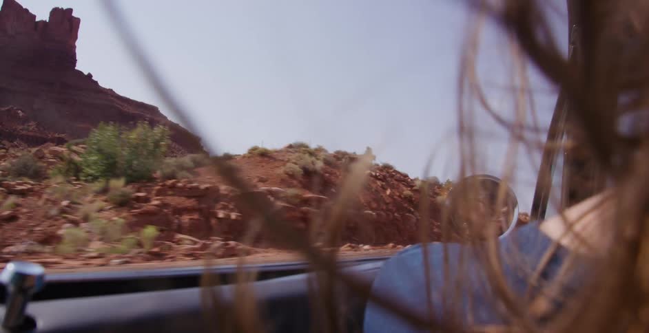 A woman's hair blows in the wind as she rides in a convertible through a desert canyon, her reflection visible in the side mirror