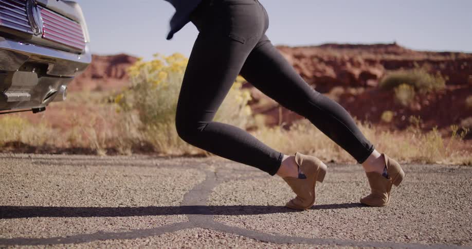 Low angle slow motion shot of a woman's legs as she struggles to push a broken-down classic car on a desert road
