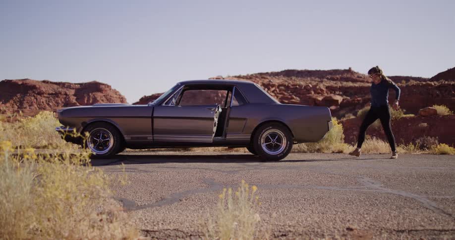 A woman struggles to push her broken down classic car down a winding desert road with red rock formations in the background