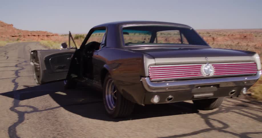 A woman gets into a classic Ford Mustang on a deserted desert road and drives away into the sunny landscape
