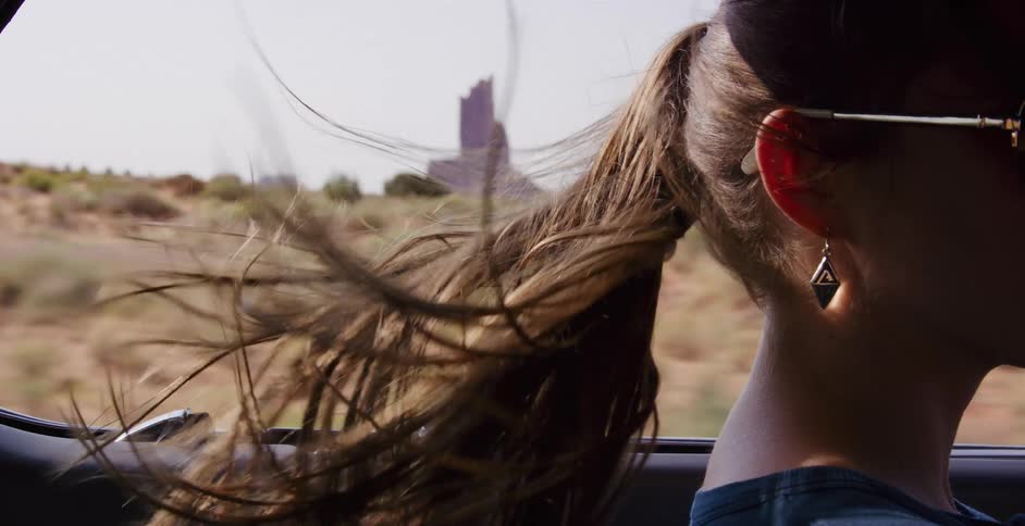 Slow motion close-up of a young woman's brown ponytail blowing wildly in the wind as she drives through a scenic desert landscape
