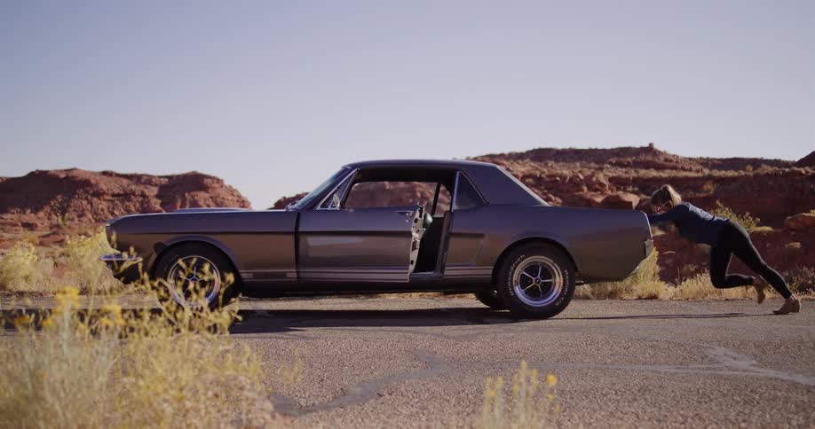 A determined woman struggles to push her broken-down vintage car on a desolate road in the sunny desert
