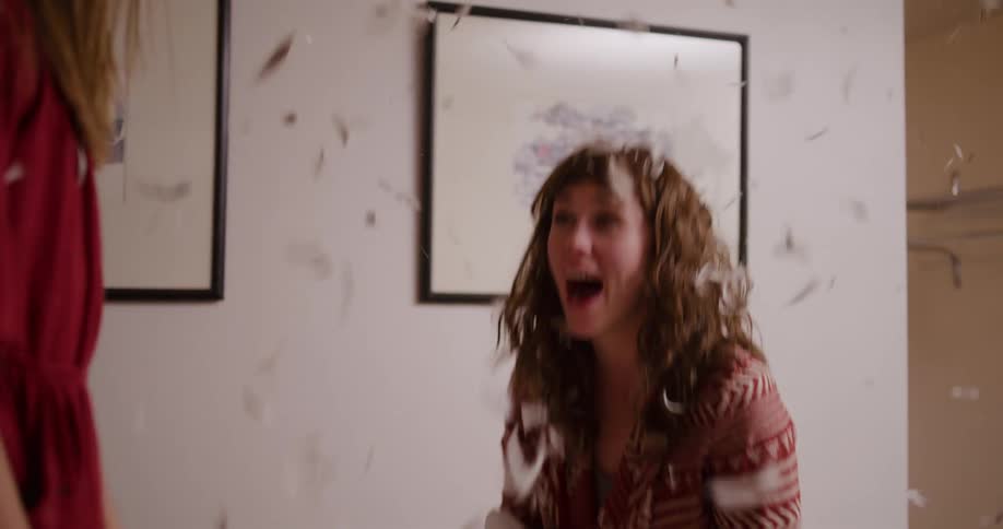 A woman laughs with joy as a pillow explodes, sending feathers flying everywhere during a playful pillow fight in slow motion