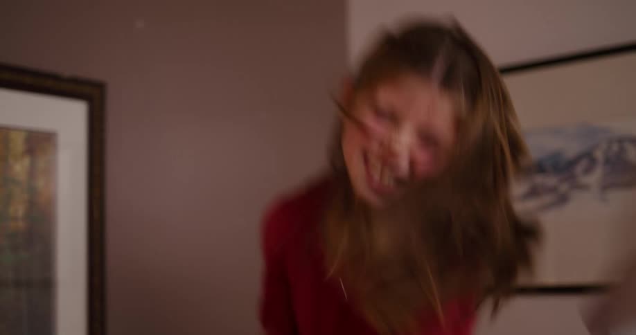 A laughing young girl in a red shirt has a pillow fight in slow motion, jumping on a bed as feathers fly all around