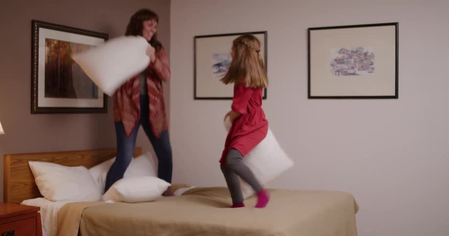 A mother and daughter joyfully jump on a bed while having a playful pillow fight in slow motion
