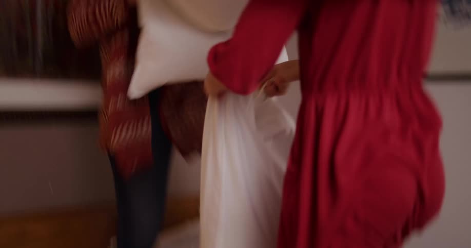 A woman and a young girl have a playful pillow fight in slow motion, laughing as white feathers fly everywhere in the bedroom