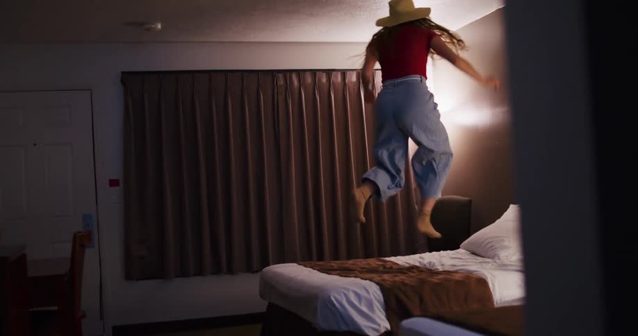A carefree young woman wearing a hat joyfully jumps up and down on a bed in a hotel room in slow motion