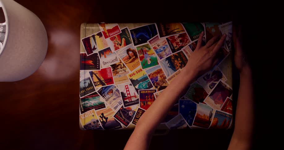 An overhead time-lapse shows hands meticulously covering a vintage suitcase with a collage of colorful travel stickers