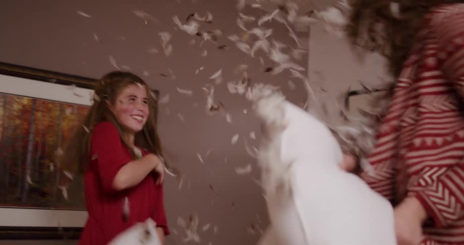 A joyful mother and daughter have a chaotic pillow fight in slow motion, with feathers flying through the air