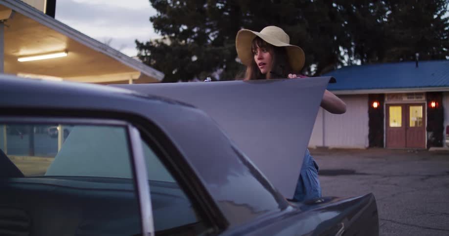 A stylish woman in a hat retrieves a vintage suitcase from the trunk of her classic car at a roadside motel and walks away