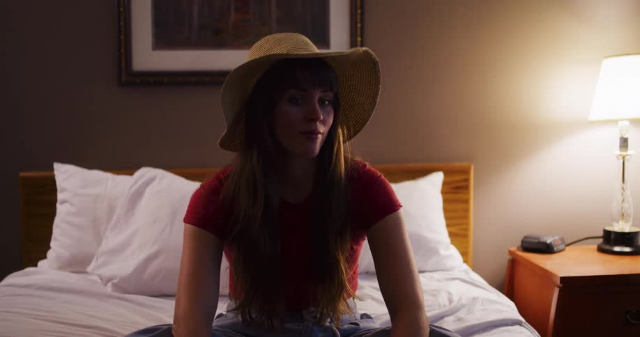 A pensive young woman wearing a sun hat sits cross-legged on a bed in a dimly lit hotel room