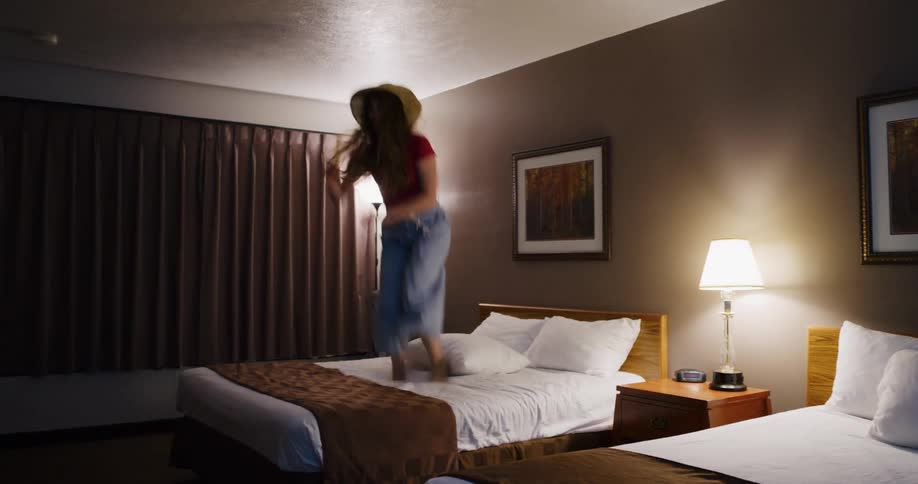A happy young woman in a wide-brimmed hat joyfully jumps on a hotel bed in slow motion