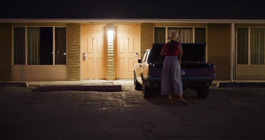 A stylish woman unloads a vintage suitcase from her classic car and walks towards her room at a roadside motel at night