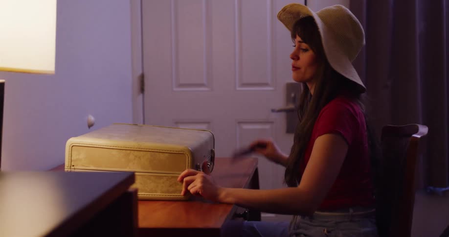 A woman in a sun hat places her vintage suitcase on a desk in a room and thoughtfully puts a postcard on top in slow motion