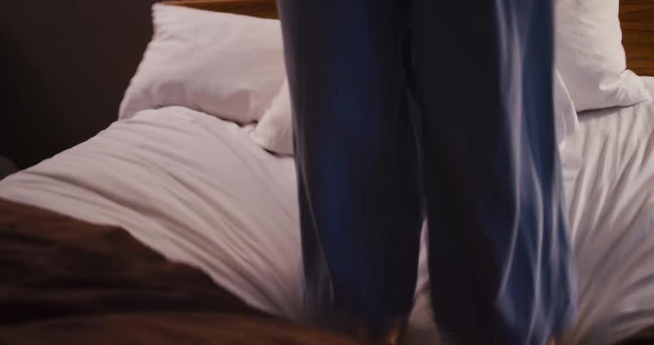 A low-angle slow-motion shot captures a happy young woman with a hat jumping with abandon on a hotel room bed