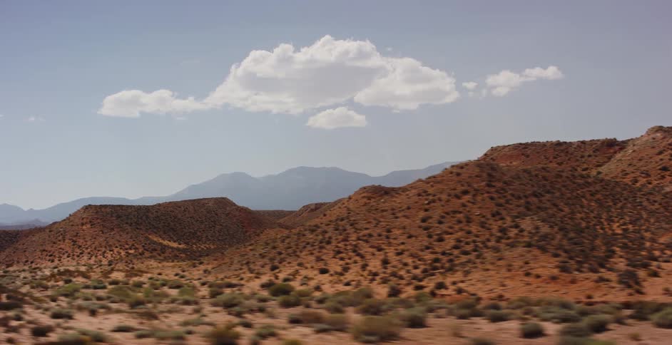 A scenic tracking shot passes by rolling red desert hills and mountains under a bright blue sky with a single white cloud