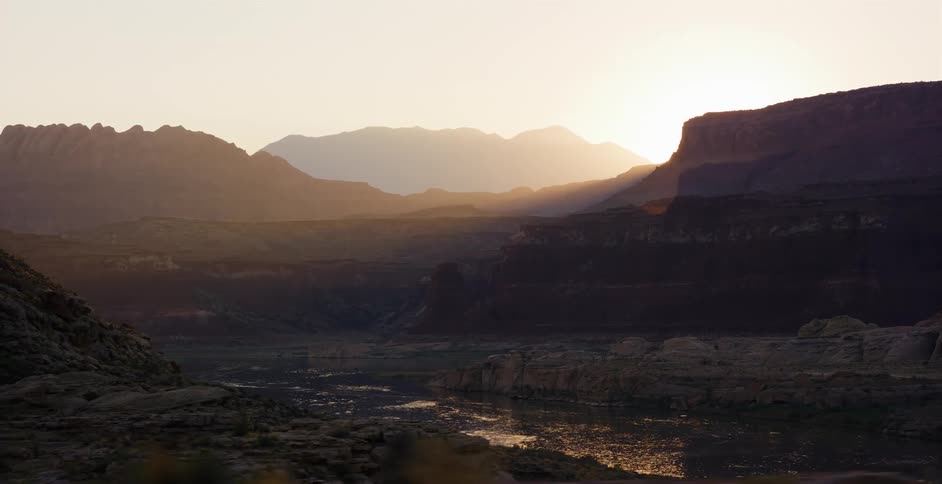 Golden sunlight washes over a vast canyon and tranquil river as the sun rises behind layered mountains