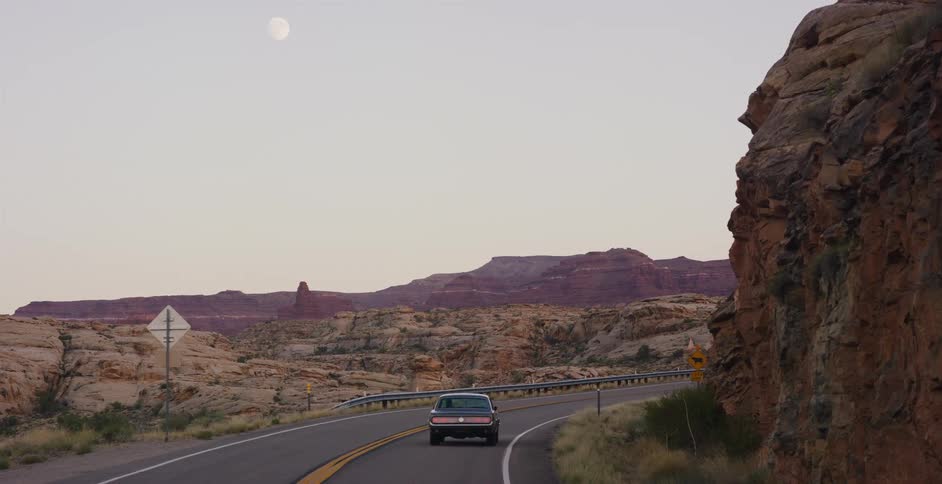 A classic car drives through a winding desert canyon highway under a full moon at dusk