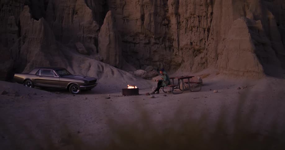 A man camps alone in a desert canyon, sitting by a campfire next to his vintage car at dusk