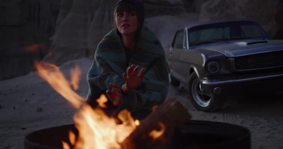 A young woman wrapped in a blanket warms her hands by a barrel fire in the desert badlands, with her classic car parked behind her