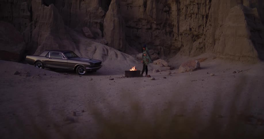 A lone traveler stands by a campfire next to their classic car in a desert canyon at twilight