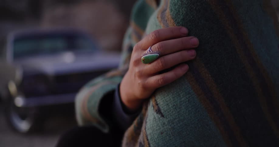 A person's hand with a silver and turquoise ring rests on their shoulder, wrapped in a warm striped blanket at dusk with a vintage car in the background