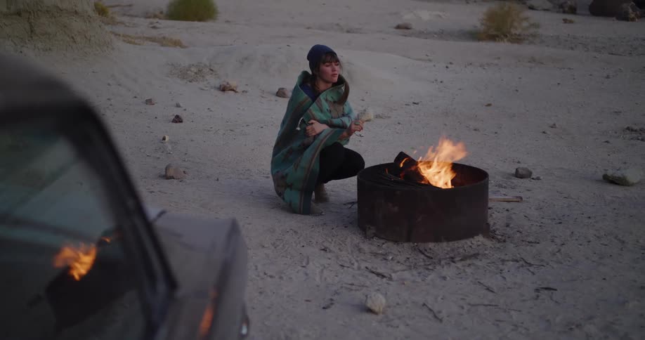 A woman wrapped in a blanket crouches by a campfire in a desert landscape next to her car at dusk