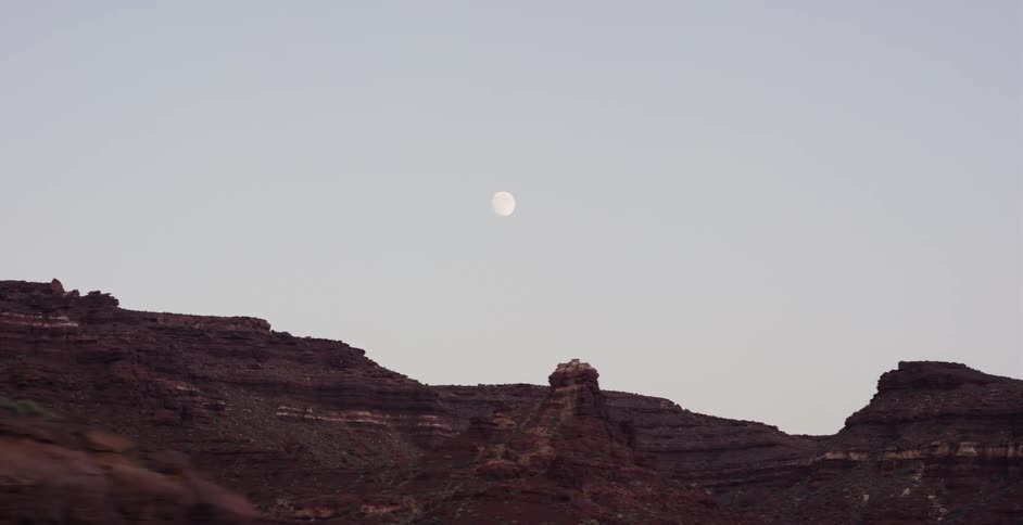 A smooth tracking shot reveals the vast red rock canyon landscape under a bright, full moon in the twilight sky