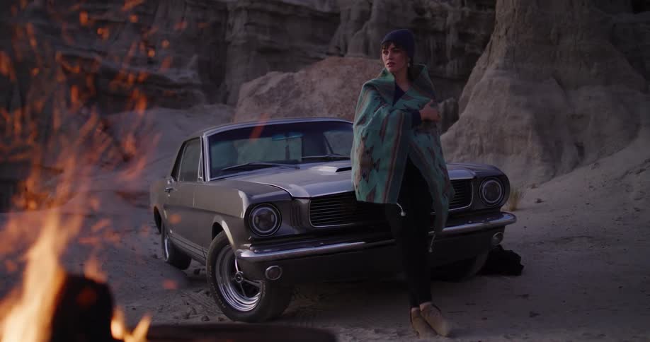 An adventurous woman wrapped in a blanket leans against her classic car next to a campfire in a desolate desert canyon at dusk