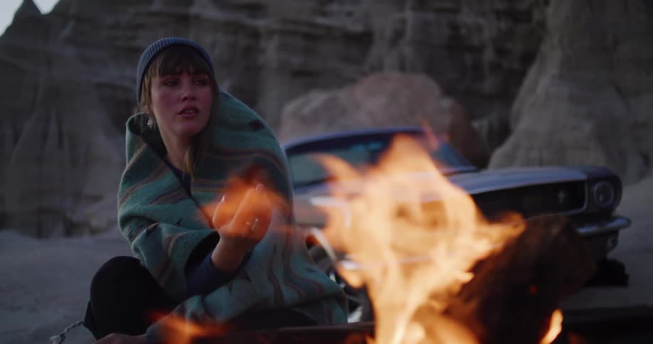 A solitary woman wrapped in a blanket warms herself by a crackling campfire in a desert canyon with her classic car in the background