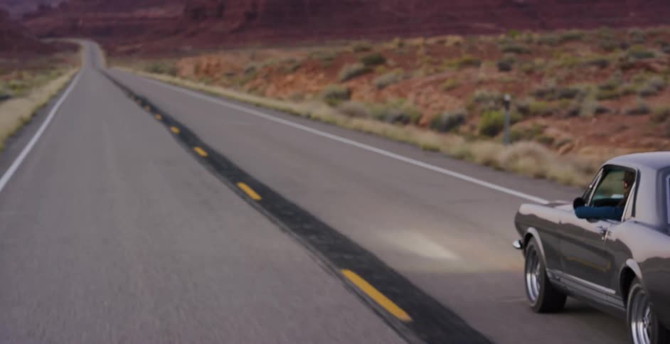 A classic grey Ford Mustang cruises down a long, empty highway through the desert at dusk
