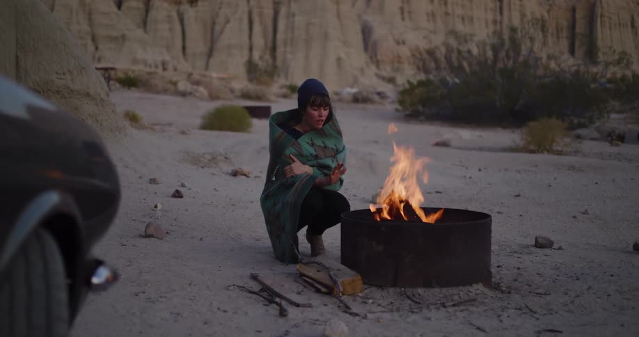 A young woman wrapped in a cozy blanket warms her hands by a campfire at a desert campsite at dusk