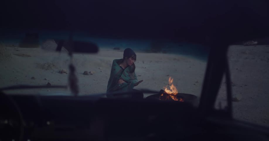 A solitary woman in a beanie and blanket sits by a crackling fire in a desolate landscape at night, seen through a car windshield