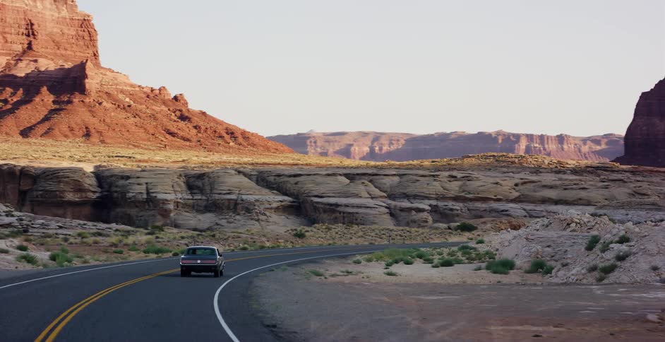 A vintage sedan navigates a winding desert highway, surrounded by majestic red rock canyons and buttes during golden hour