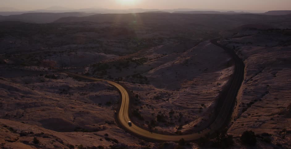 An aerial view captures a lone car navigating a serpentine desert highway as the sun sets over the vast, empty landscape