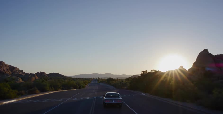 A vintage muscle car drives down an empty desert highway into the beautiful, cinematic sunset
